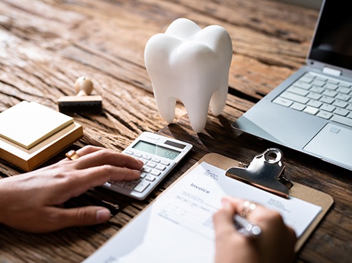 Someone calculating dental invoice at a desk with calculator, laptop, and large model tooth